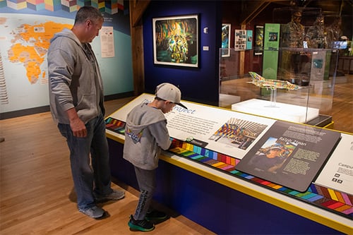 Young boy in ball cap with guardian touching beaded panel in The MiG-21 Project exhibit